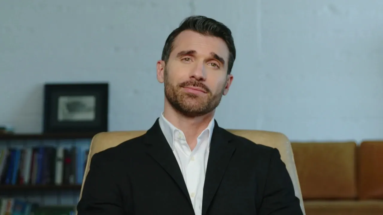 Dr Ed AI sex educator avatar: a bearded man in a black blazer and white shirt seated in a tan chair, bookshelf behind him, ready for a private video chat